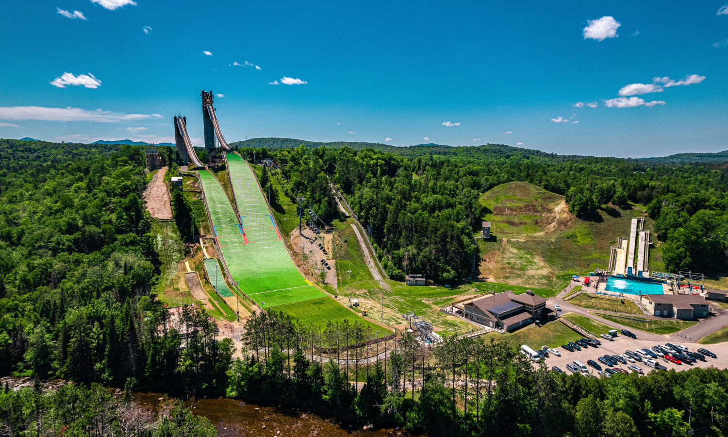 Ski jump with green hill, buildings, trees, and blue sky.