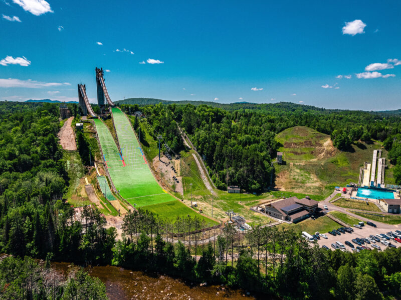 Ski jump with green hill, buildings, trees, and blue sky.