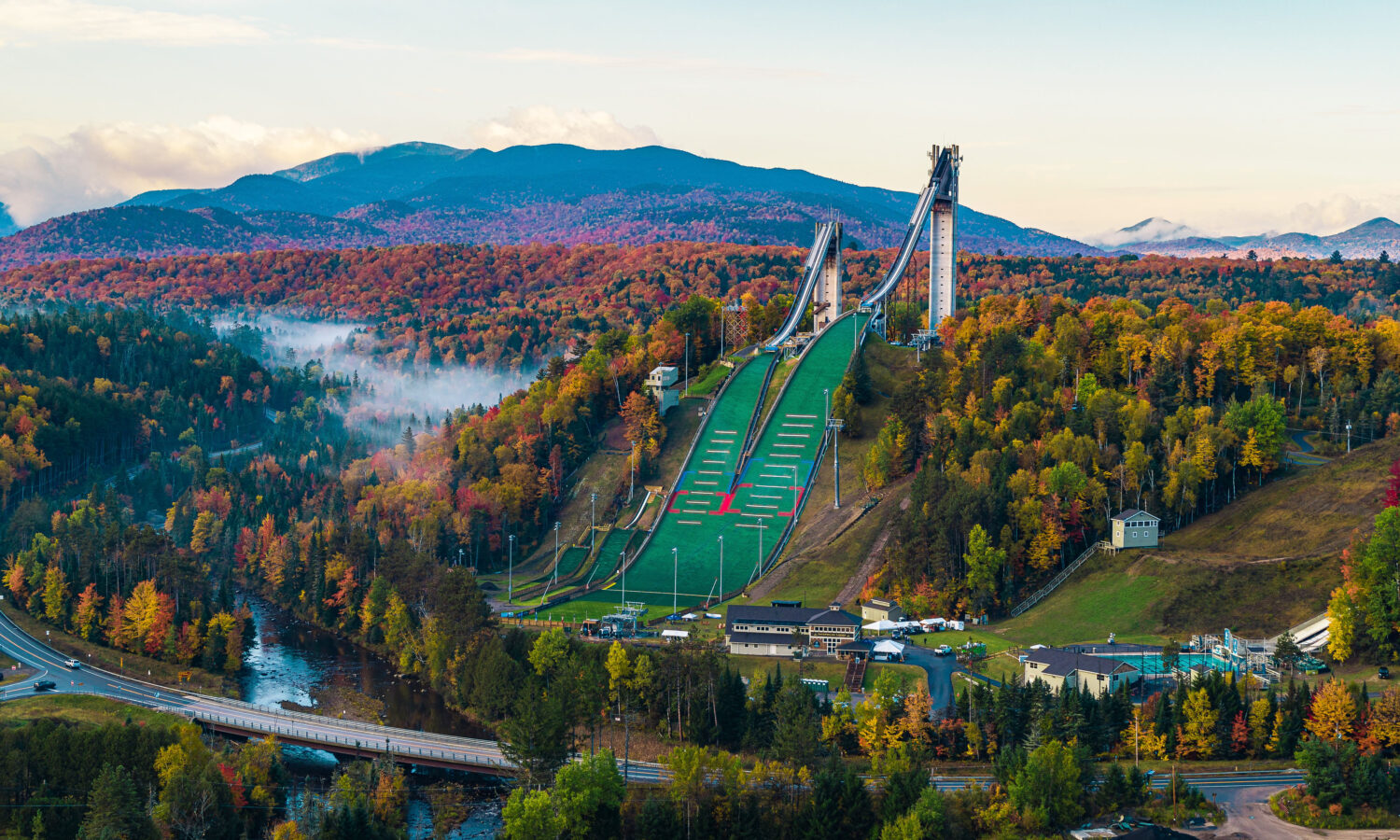 Ski jumps on green hill amid autumn trees and mountains.