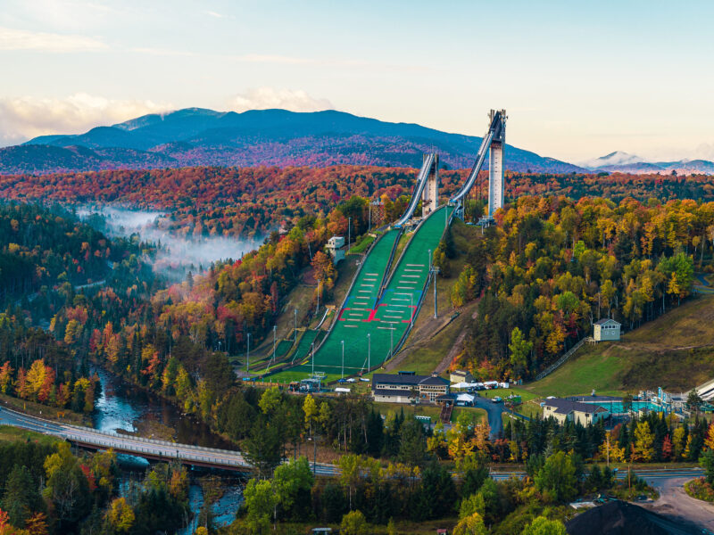 Ski jumps on green hill amid autumn trees and mountains.