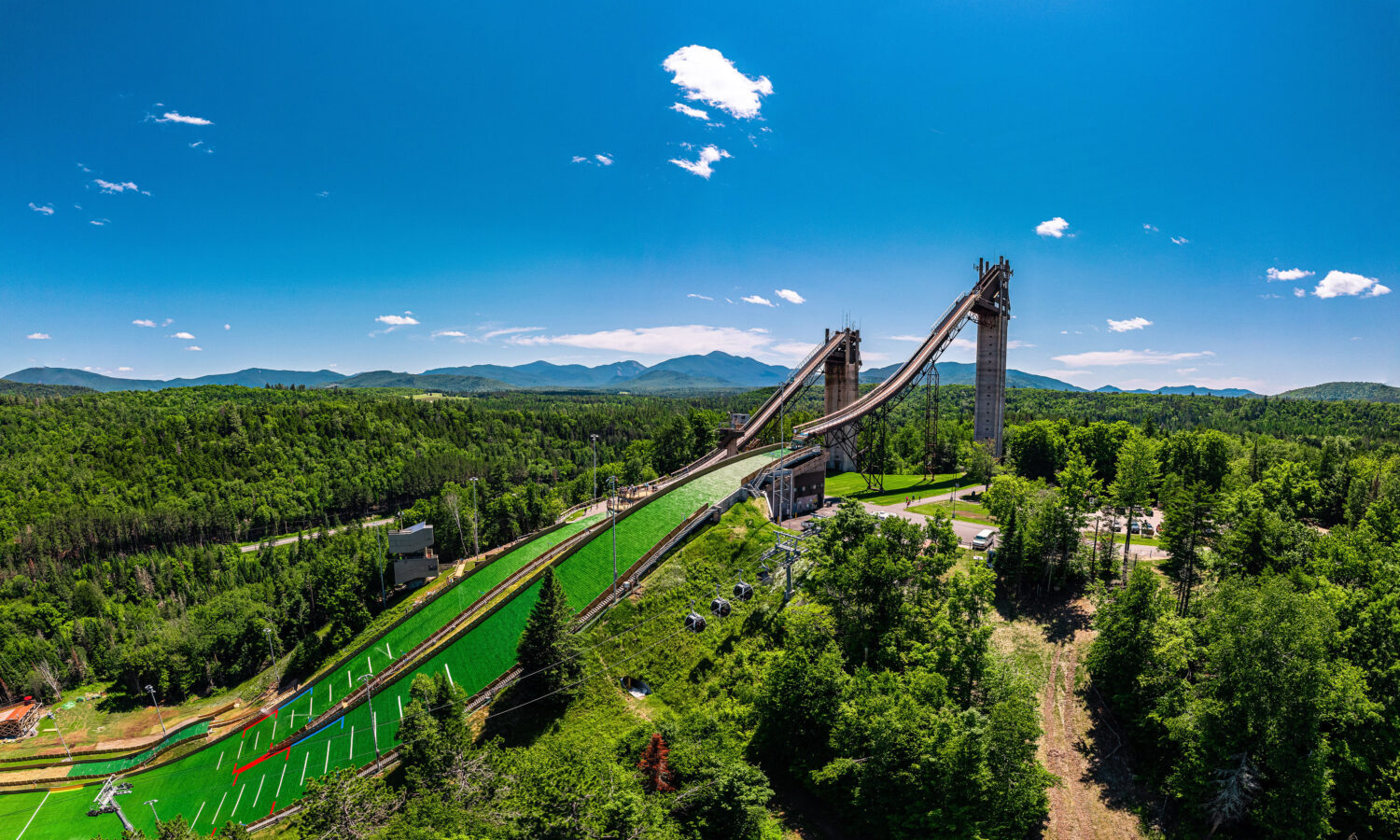 Ski jumps amid forest, with mountains and clear blue sky.