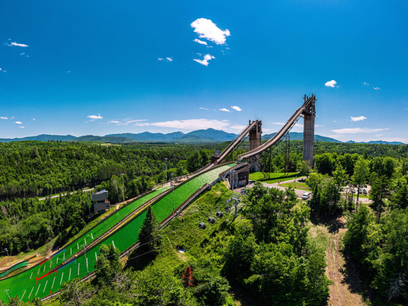 Ski jumps amid forest, with mountains and clear blue sky.