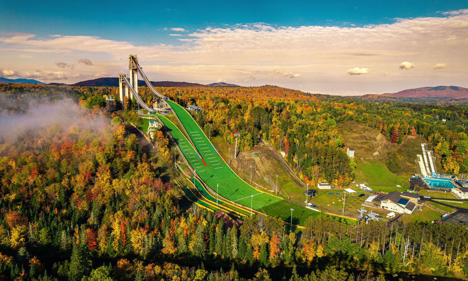 Aerial of ski jump amid colorful autumn trees, blue sky.