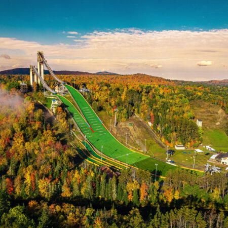 Aerial of ski jump amid colorful autumn trees, blue sky.