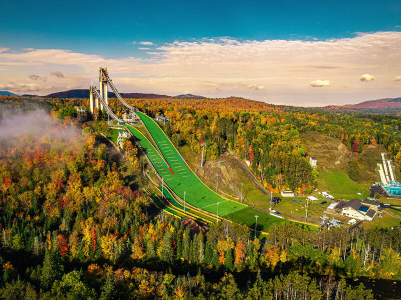 Aerial of ski jump amid colorful autumn trees, blue sky.