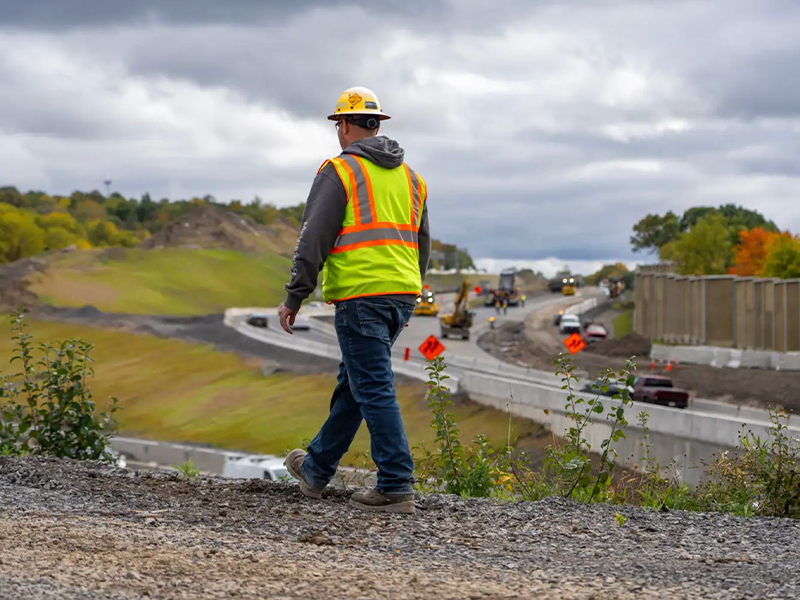 Construction worker walks on gravel near highway under cloudy sky.