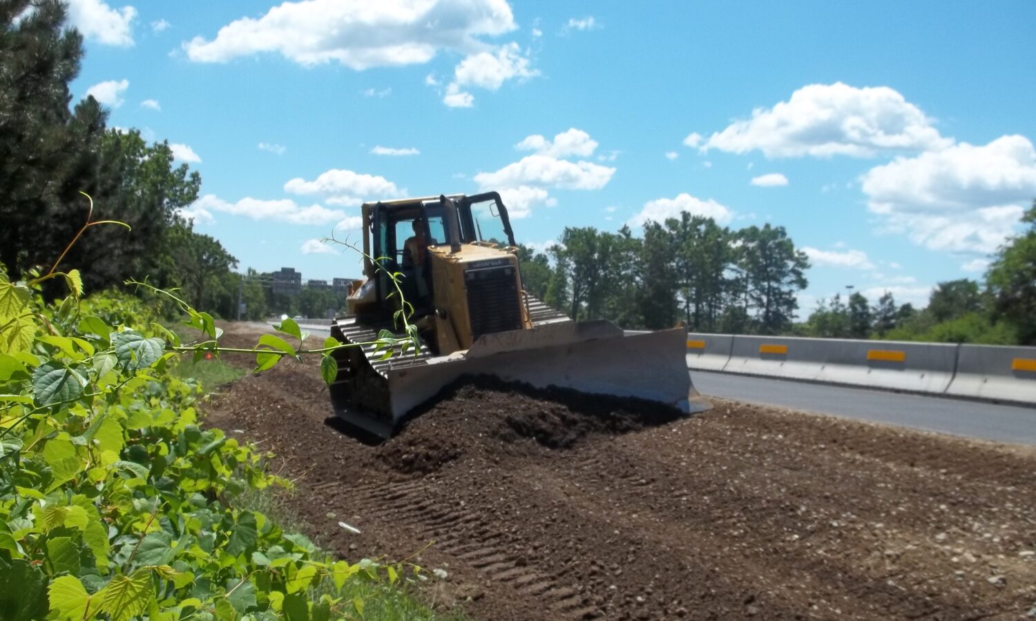 Bulldozer spreads dirt by road under partly cloudy sky.