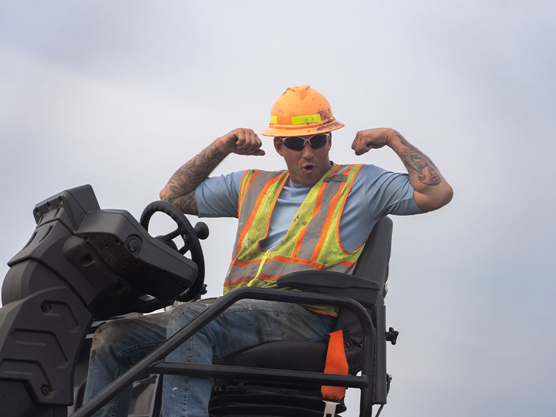 Worker in hard hat flexes biceps while sitting on machinery.