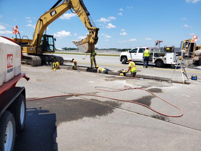 Construction workers repair pavement beside machinery on a clear day.