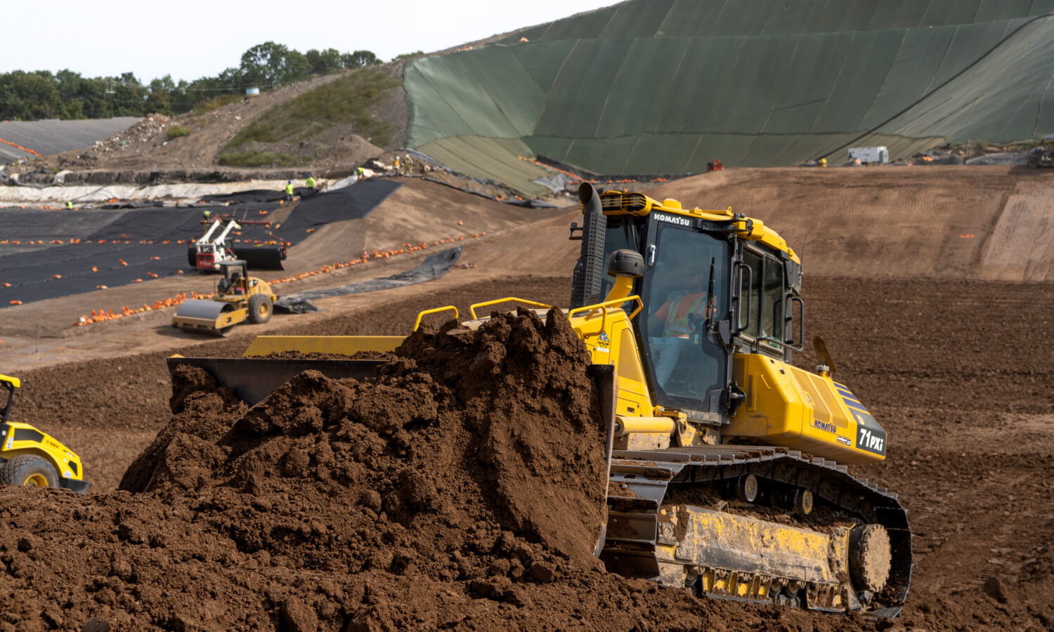 Yellow bulldozer moves soil with machinery and tarped slopes behind.