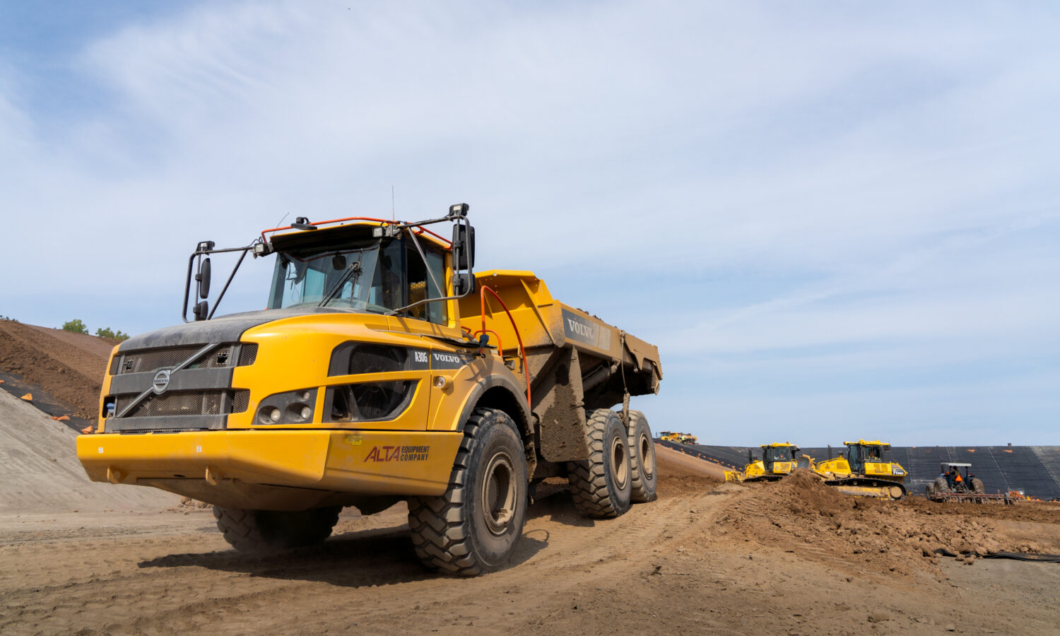Yellow Volvo dump truck drives on dirt site, others behind.