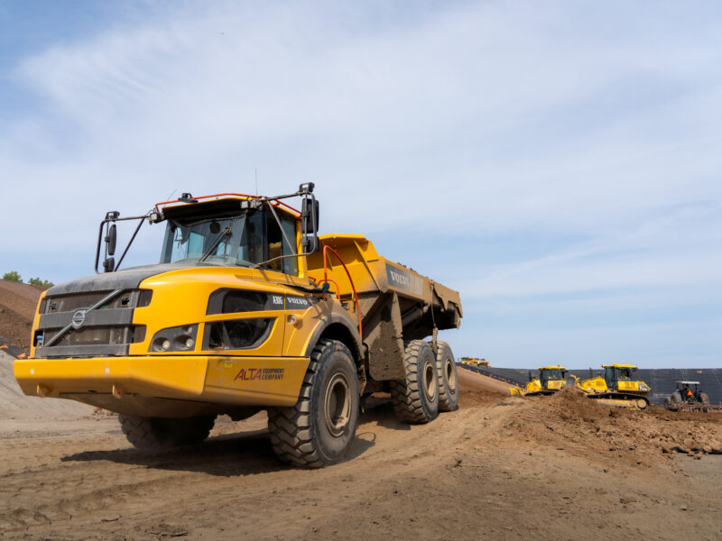 Yellow Volvo dump truck drives on dirt site, others behind.