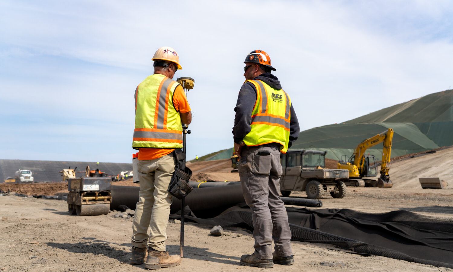 Two construction workers stand on a dirt site with surveying equipment.
