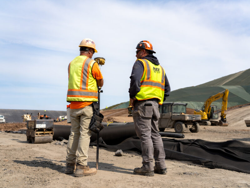 Two construction workers stand on a dirt site with surveying equipment.
