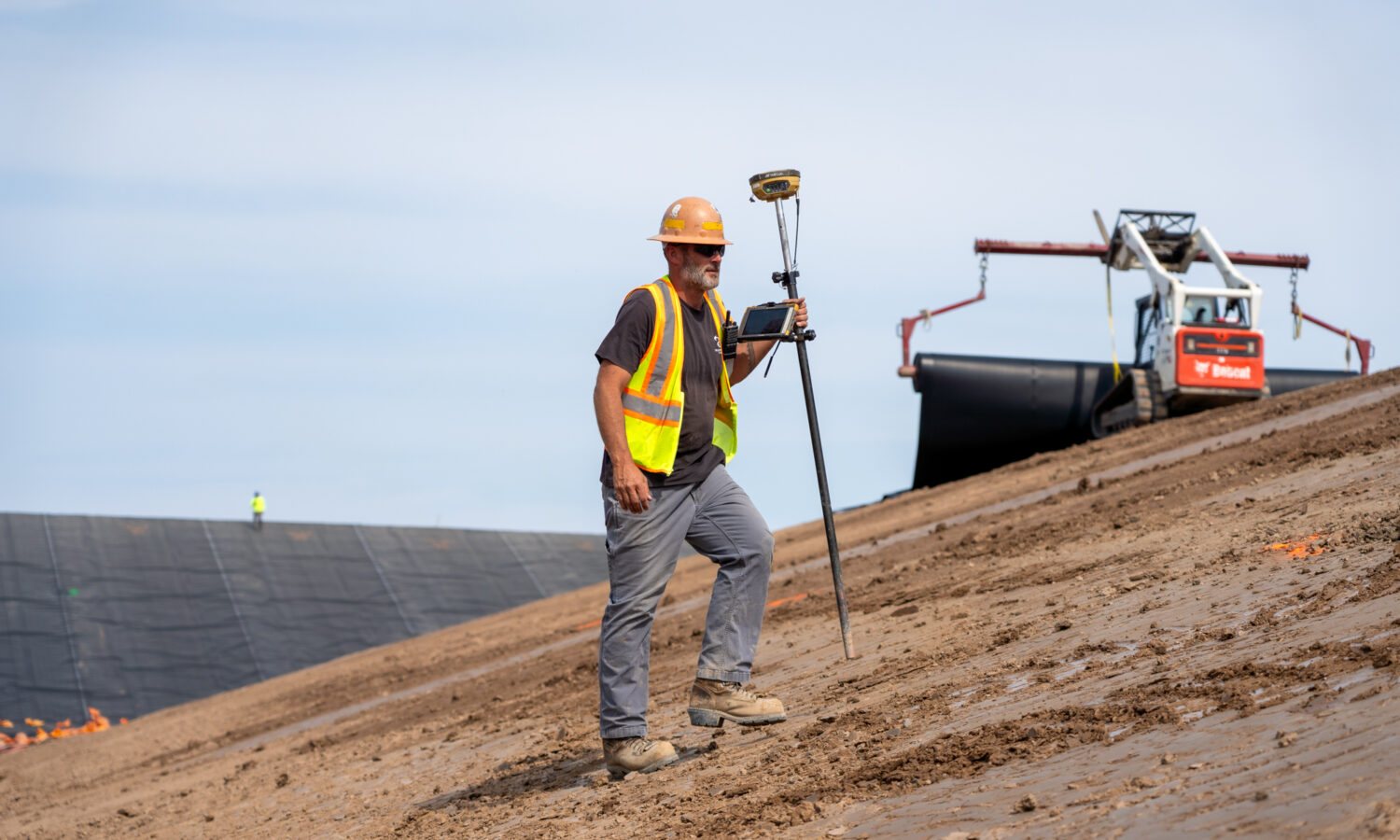 Worker in yellow vest surveys sloped site; machinery behind.