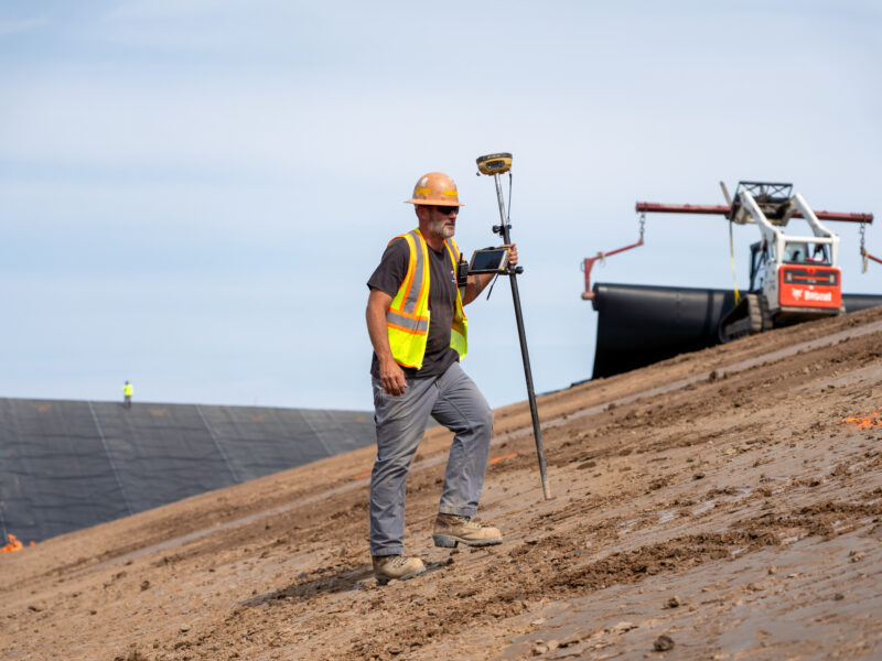 Worker in yellow vest surveys sloped site; machinery behind.