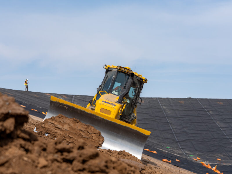 Yellow bulldozer moves dirt; worker stands by tarp-covered slope.