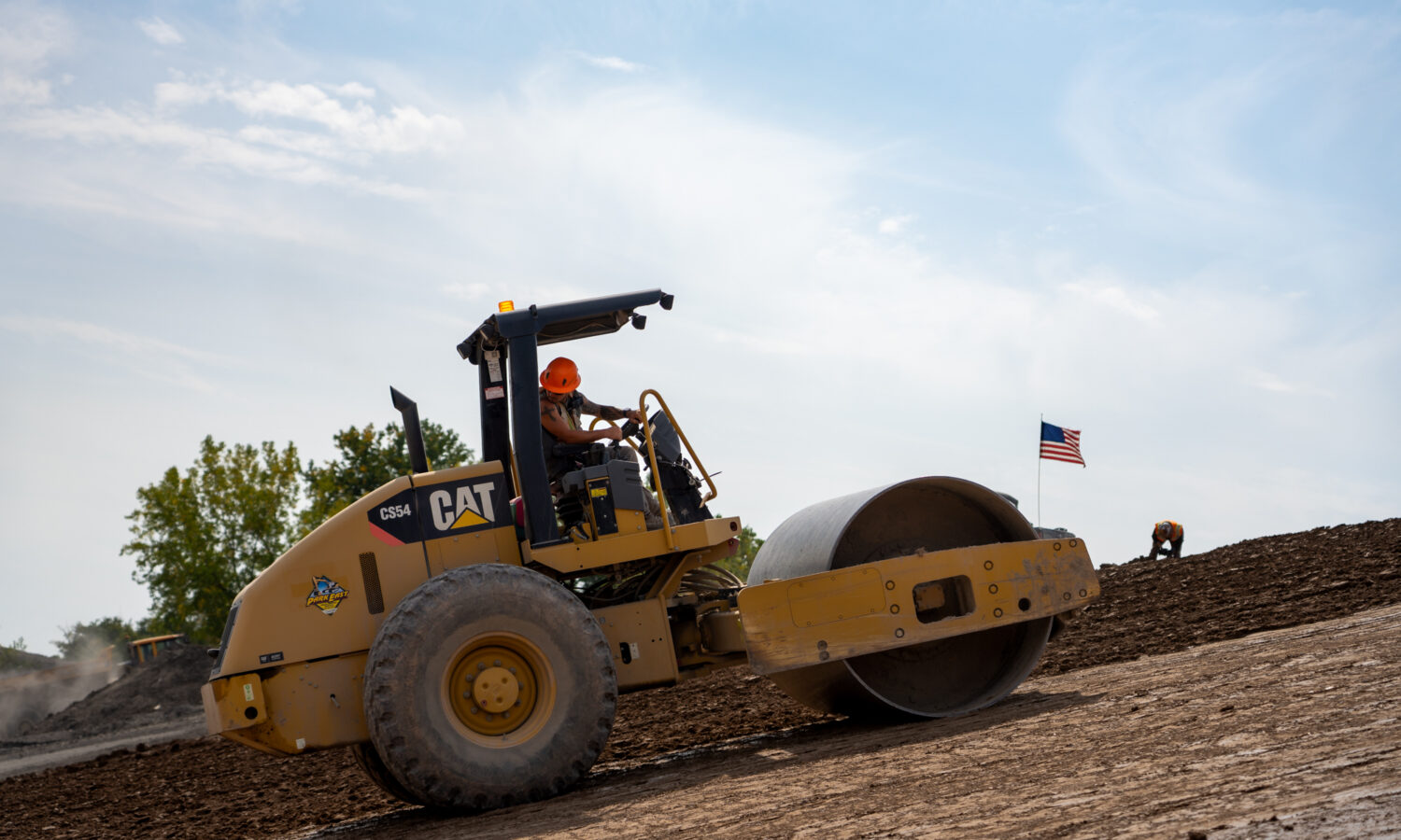 Worker drives soil compactor on slope; American flag, coworker behind.