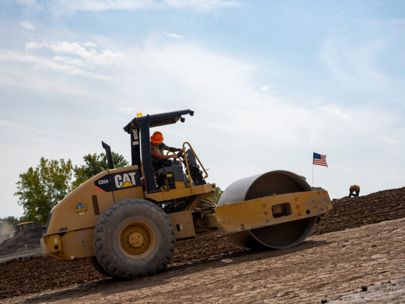 Worker drives soil compactor on slope; American flag, coworker behind.