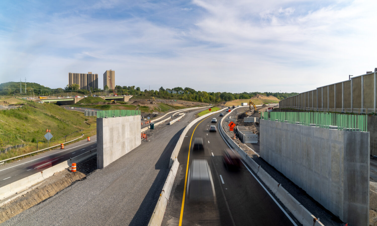 Vehicles drive on finished lanes beside barriers and construction equipment.
