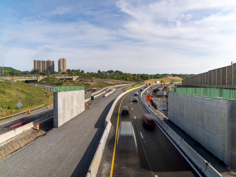 Vehicles drive on finished lanes beside barriers and construction equipment.