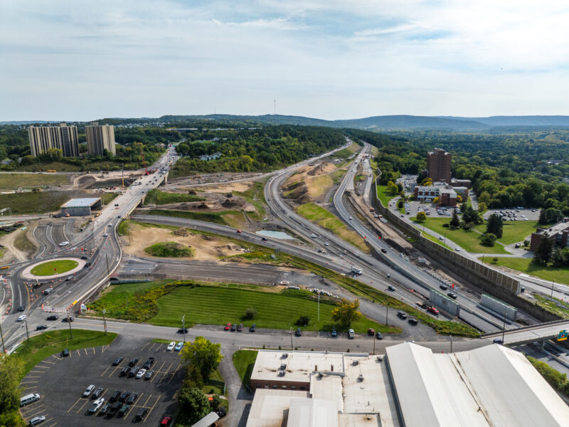 Aerial view of highway construction with nearby buildings and greenery.