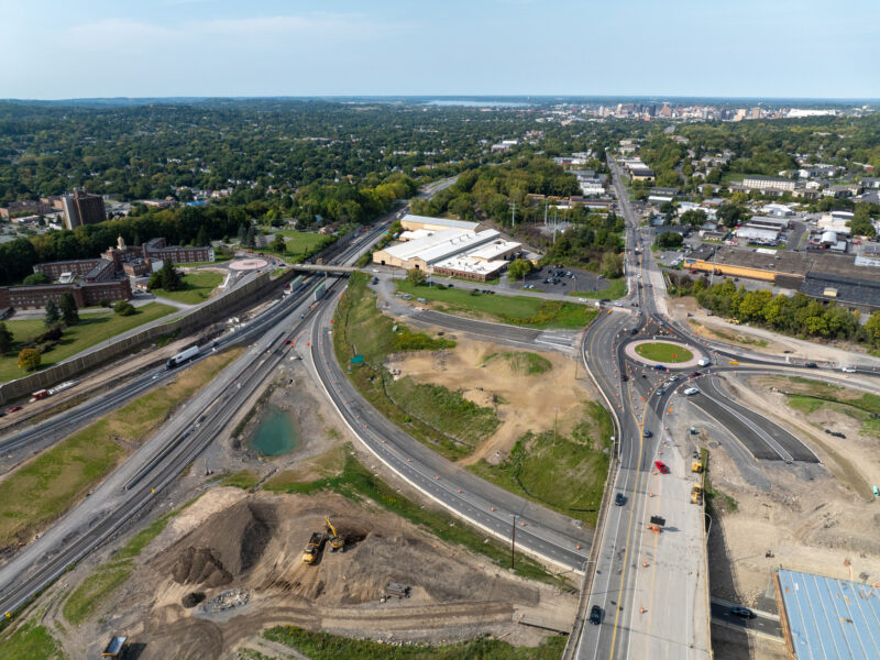 Aerial view: highway roundabout, buildings, vehicles, green spaces.