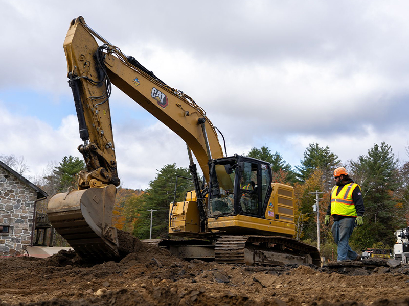 A worker stands by a yellow excavator at a construction site.