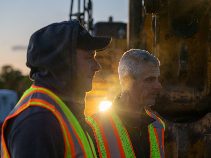 Two men in safety vests stand by construction equipment, backlit.