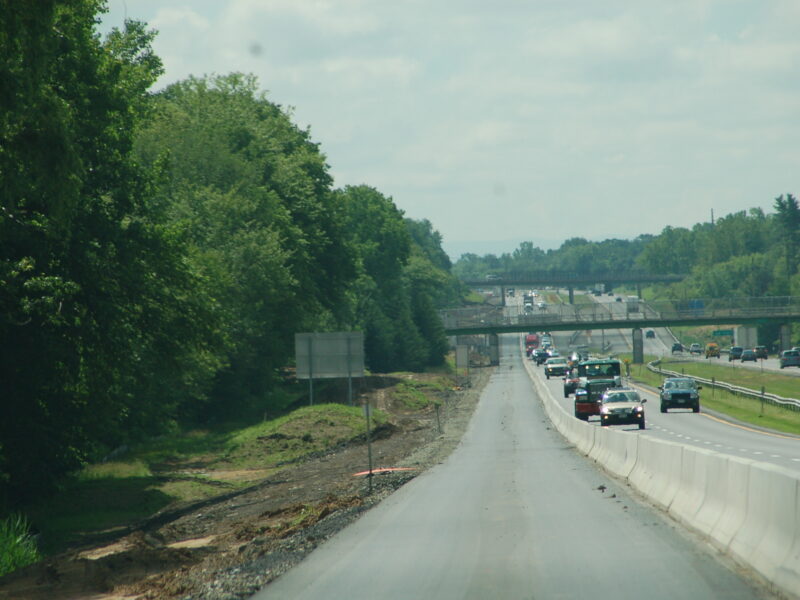 Cars drive in one lane beside barriers; trees, overpasses.