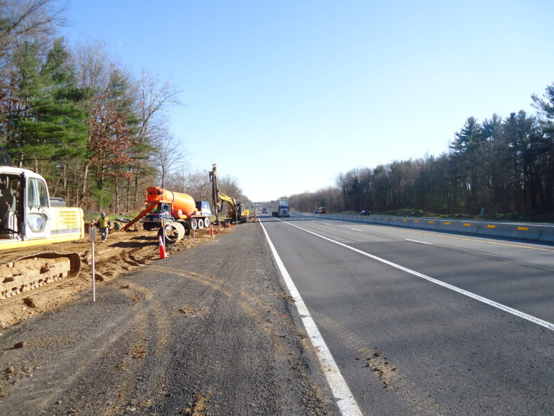 Construction vehicles line the highway behind traffic barriers under clear skies.