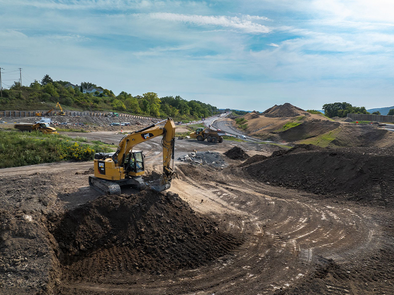 Excavator digs dirt mound at construction site near road.