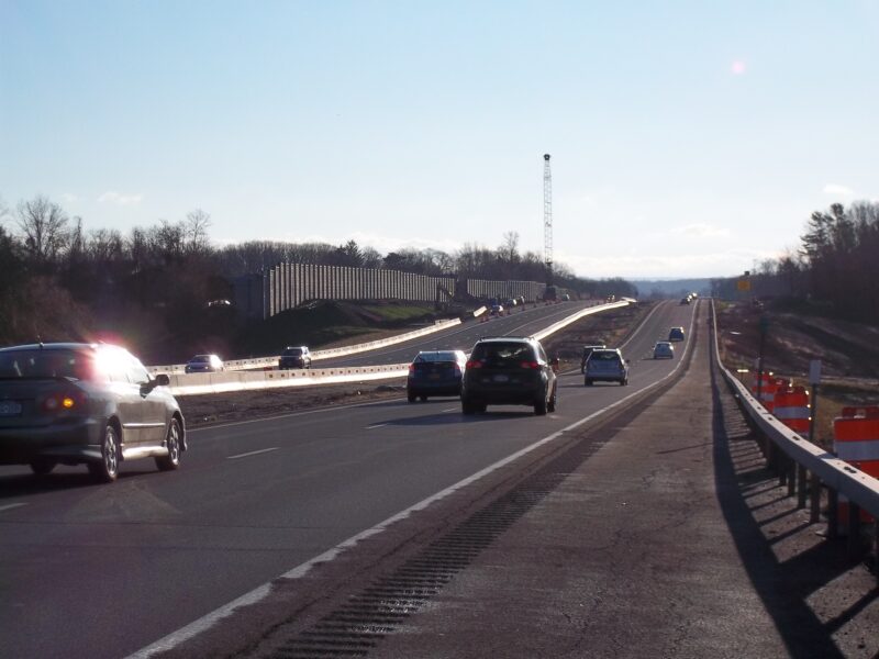 Cars on highway, sound barrier left, crane ahead, clear sky.