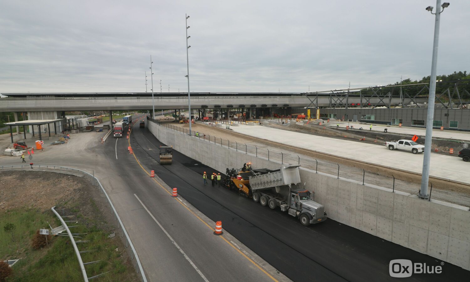 Workers and machines lay asphalt beneath overpass, barriers line road.
