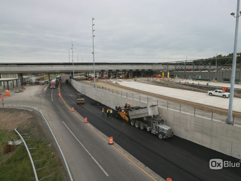 Workers and machines lay asphalt beneath overpass, barriers line road.