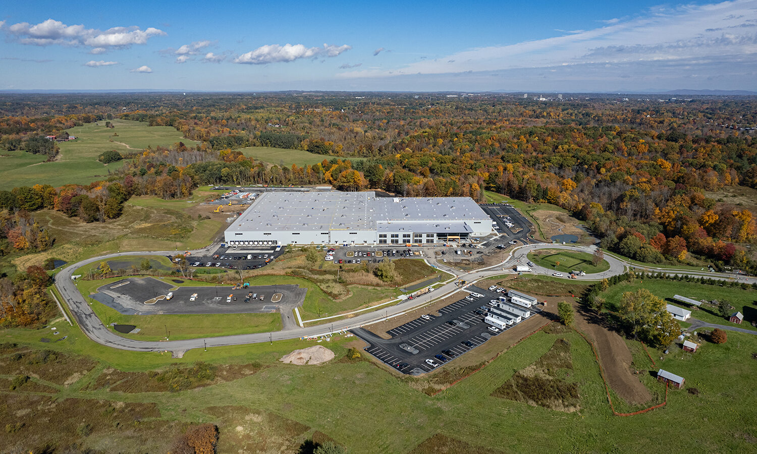Aerial of large warehouse, trucks, parking lots, forest, sky.