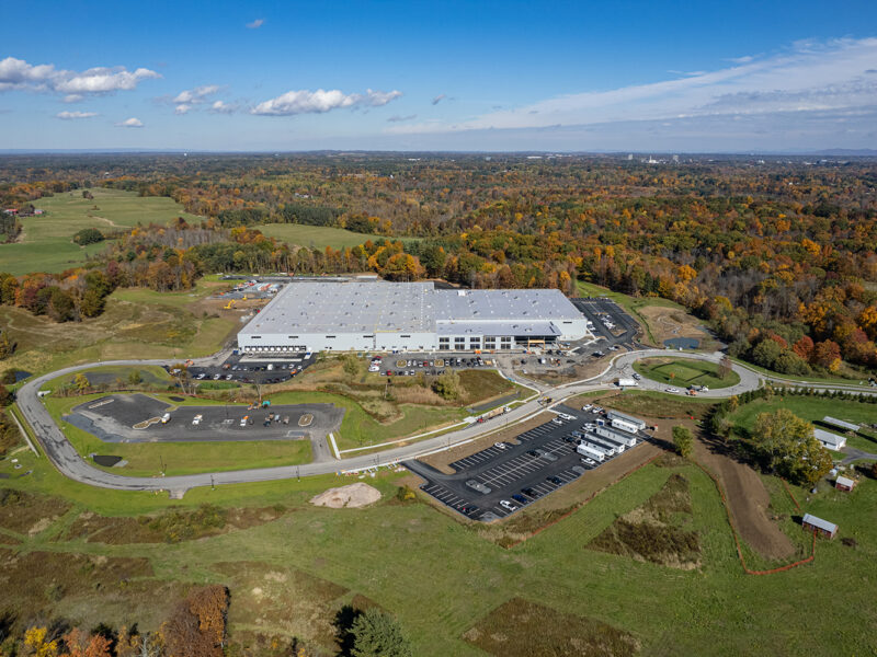 Aerial of large warehouse, trucks, parking lots, forest, sky.