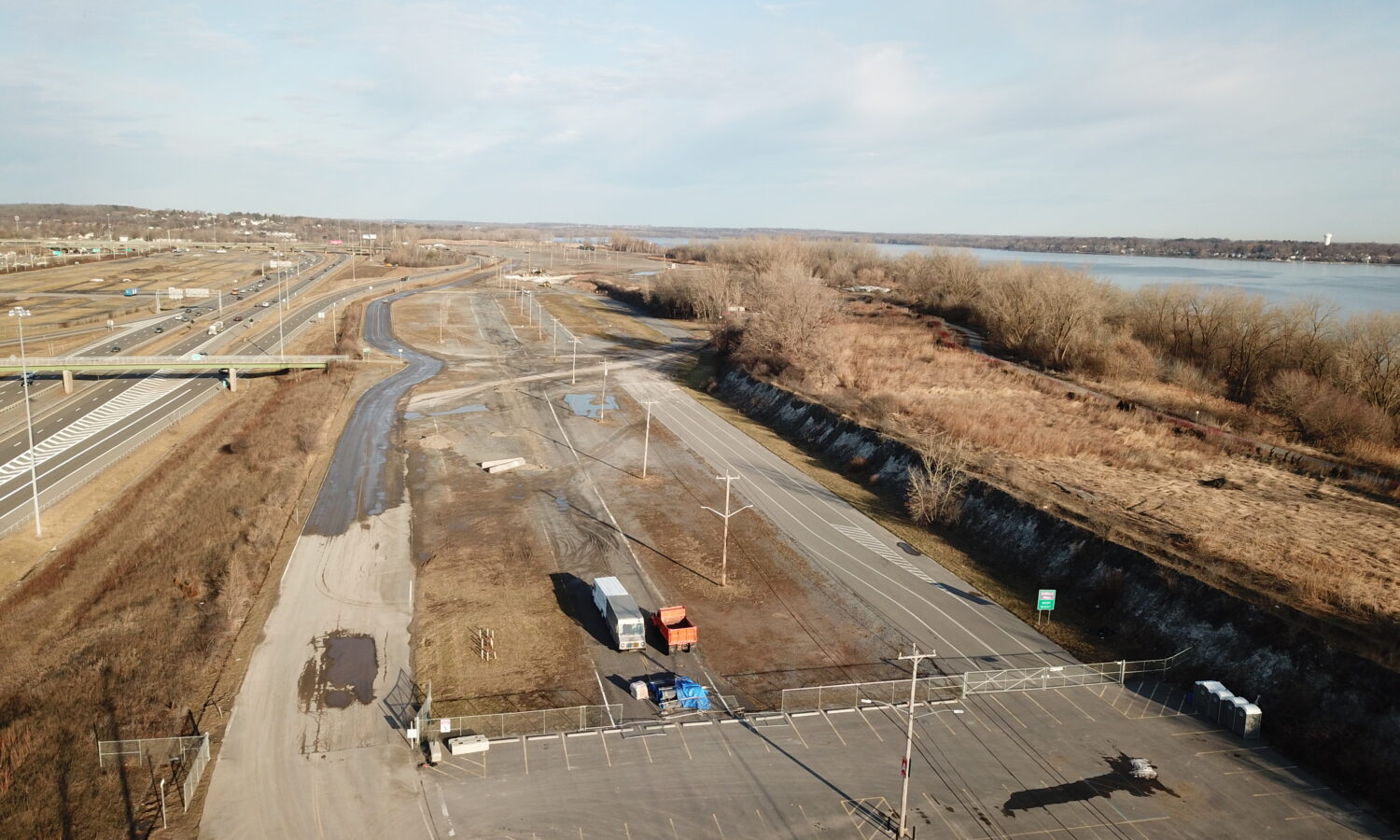Drone shot: empty fenced parking lot by river, roads, trees.