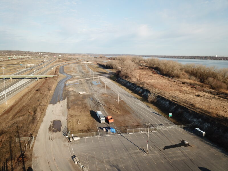Drone shot: empty fenced parking lot by river, roads, trees.