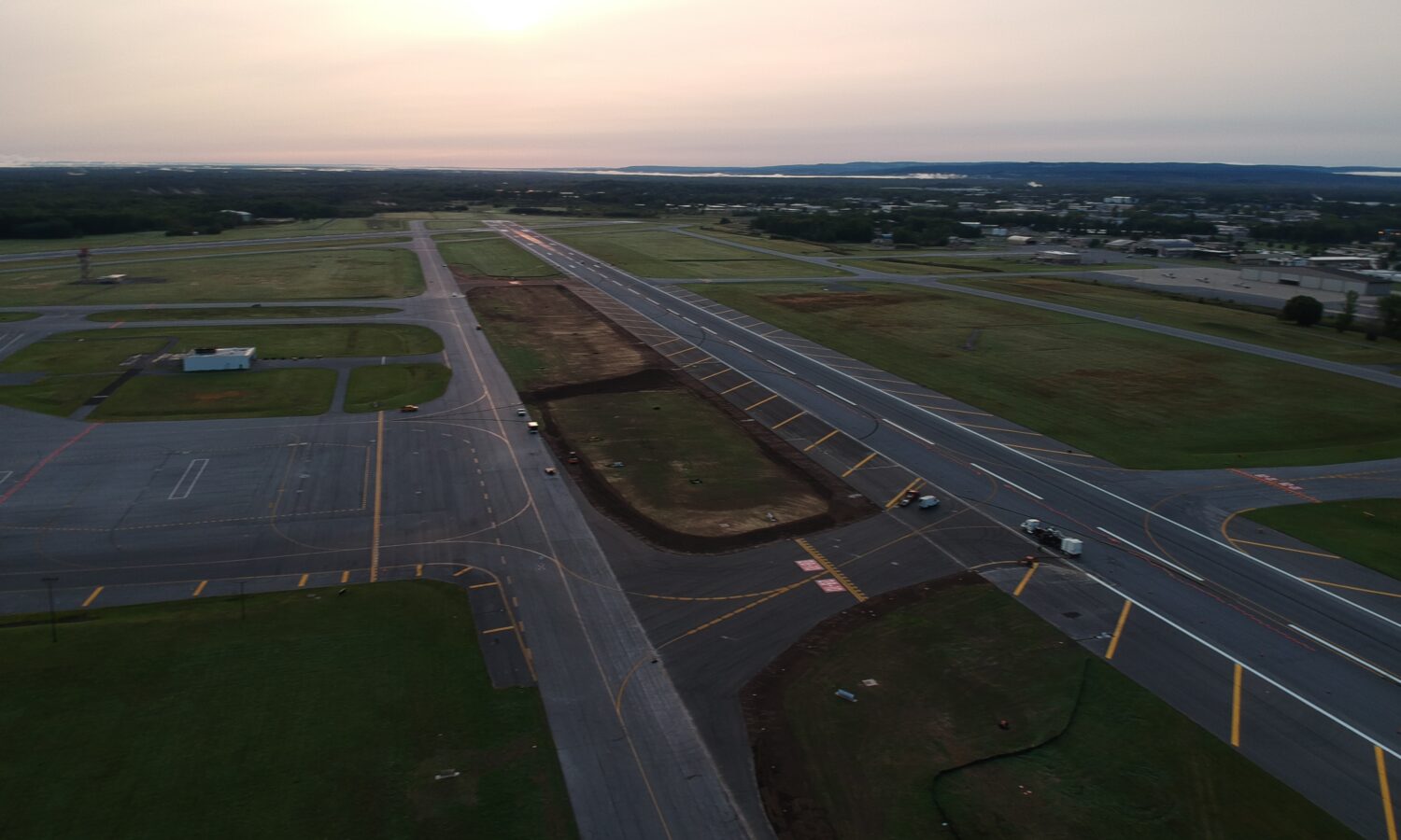 Aerial sunset view of runway construction with vehicles visible.