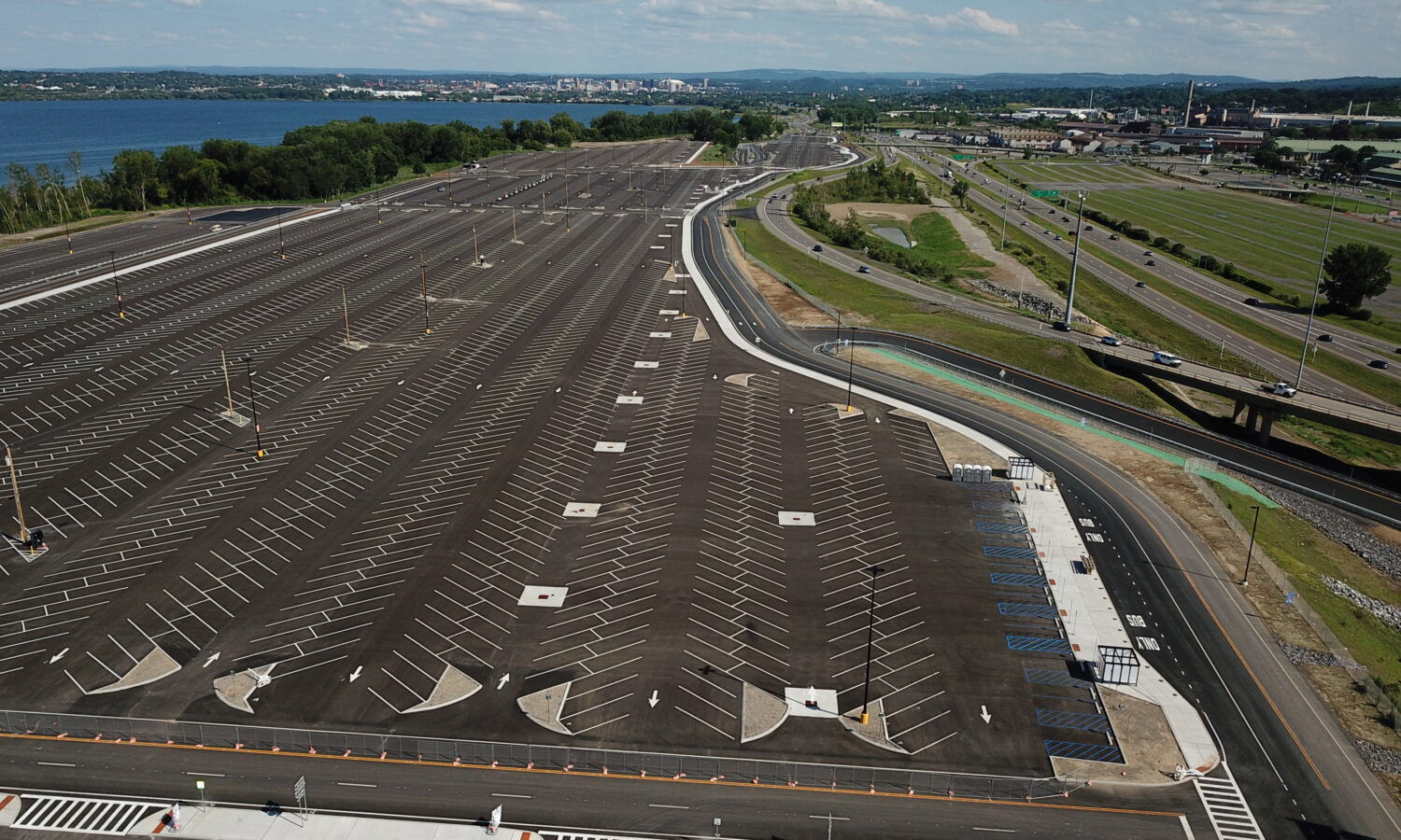 Empty parking lot by water and highway, under cloudy sky.