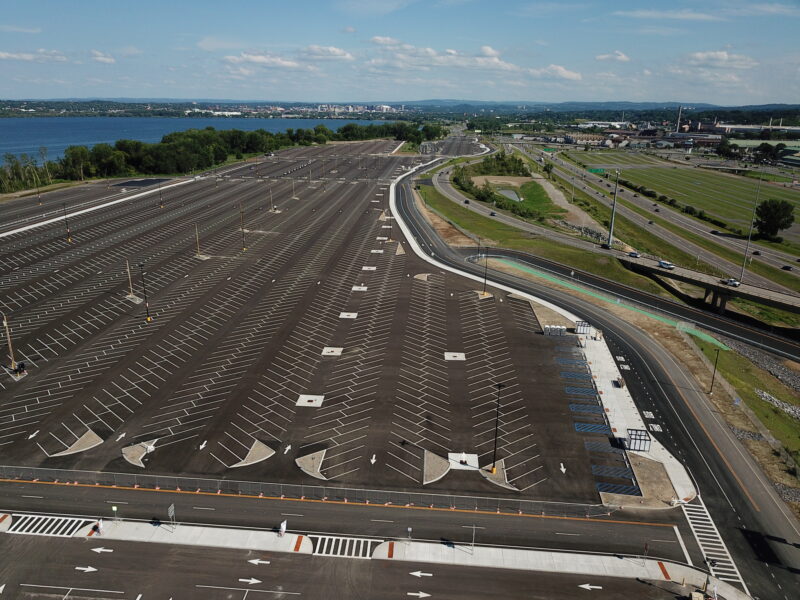 Empty parking lot by water and highway, under cloudy sky.
