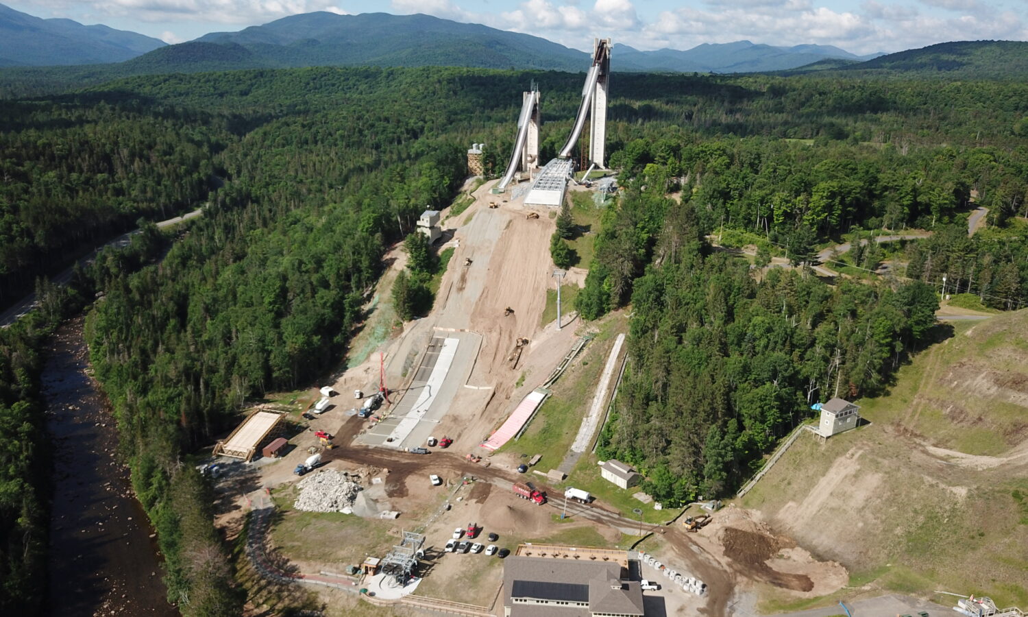 Aerial view: ski jumps, construction, pool, dense forest, mountains.