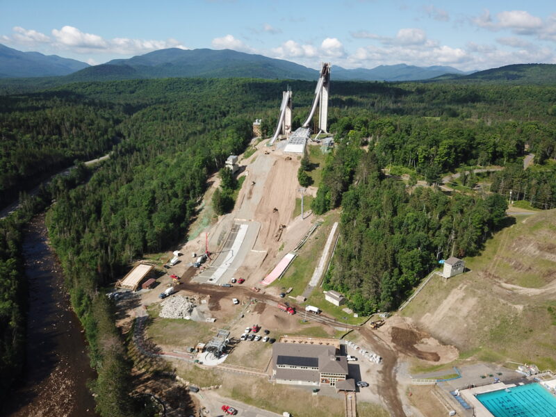 Aerial view: ski jumps, construction, pool, dense forest, mountains.