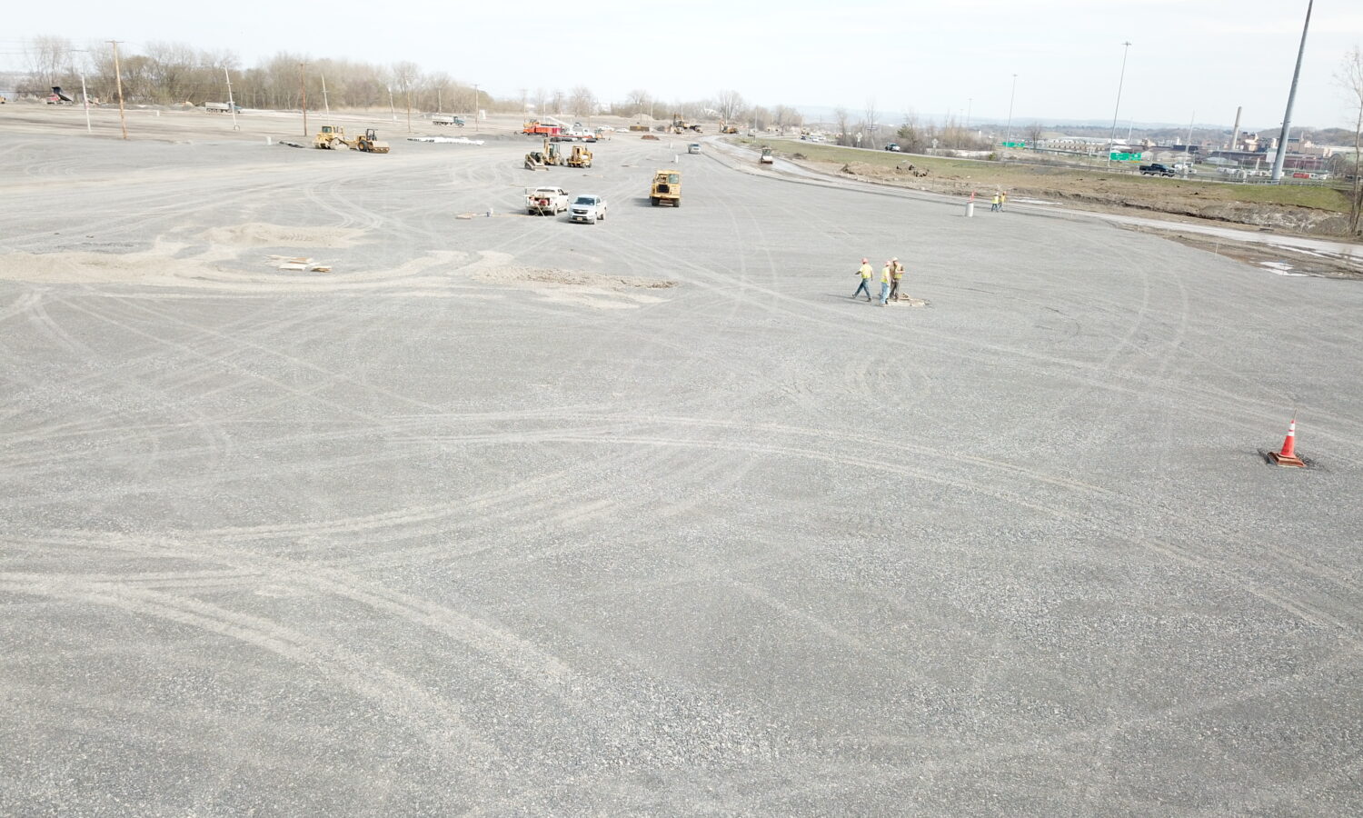 Gravel lot with scattered vehicles, workers, cones, and tire tracks.