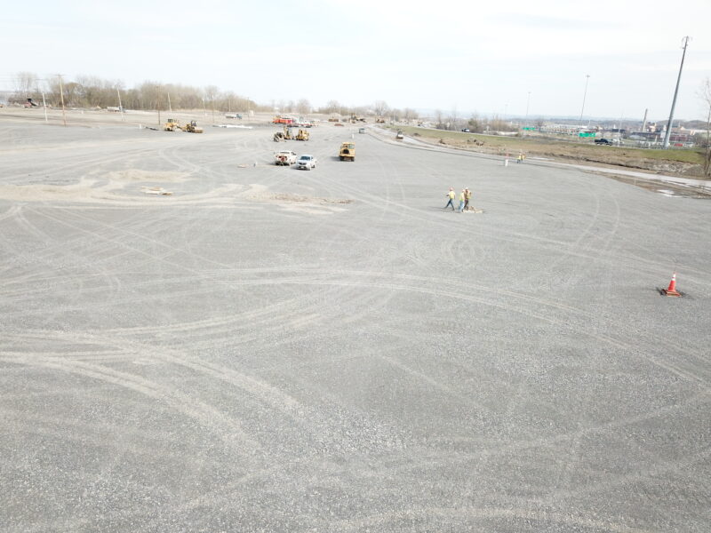 Gravel lot with scattered vehicles, workers, cones, and tire tracks.