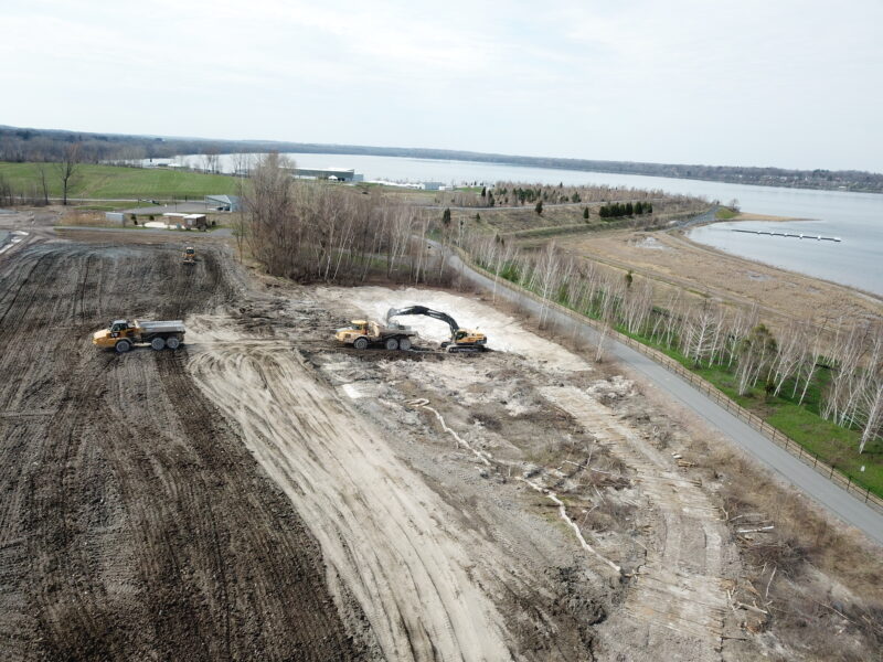 Aerial view: construction vehicles clear land by water and road.