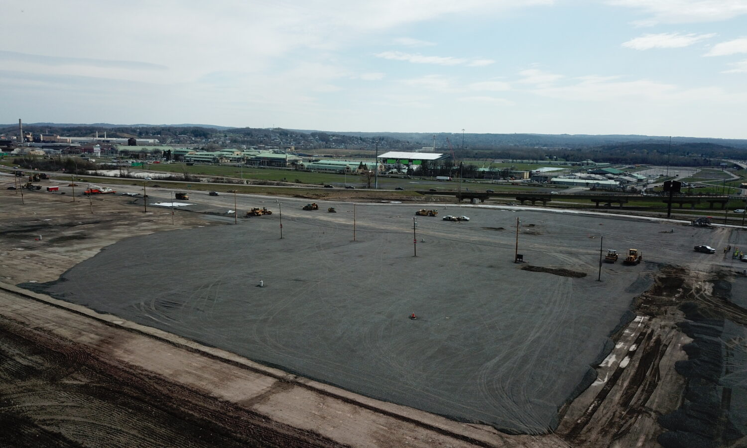 Spacious gravel site with scattered vehicles, equipment, and distant buildings.