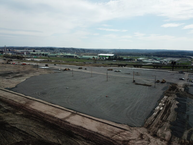 Spacious gravel site with scattered vehicles, equipment, and distant buildings.