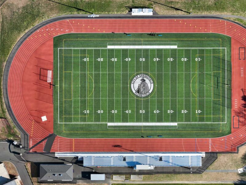 Aerial view: football field with mascot, track, bleachers, courts.